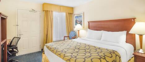 A hotel room with a bed, a desk, a chair, a lamp, and a door.