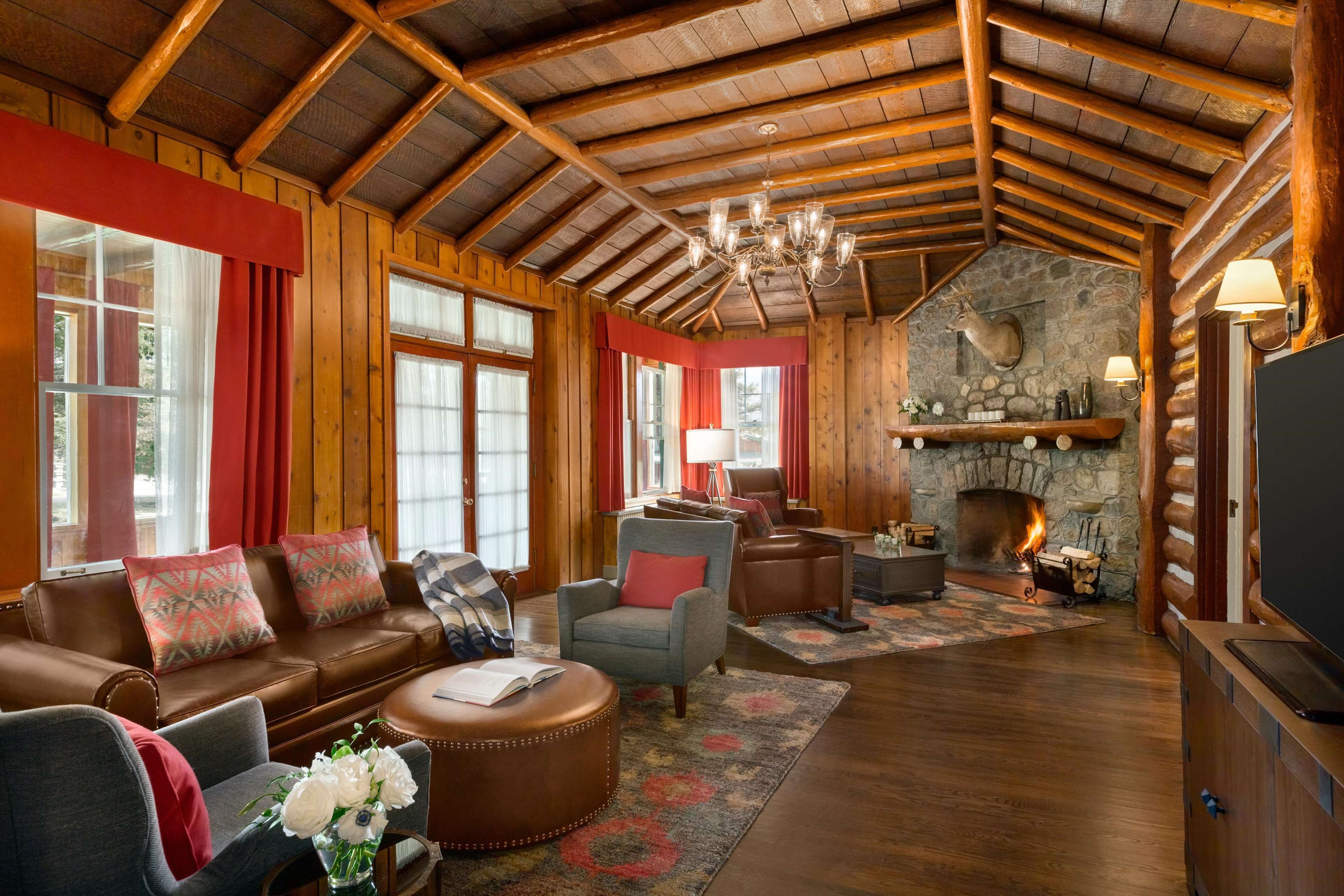 Interior de una cabaña de troncos con chimenea de piedra, vigas de madera, muebles de cuero y un televisor.