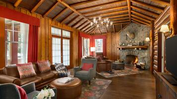 Interior de una cabaña de troncos con chimenea de piedra, vigas de madera, muebles de cuero y un televisor.