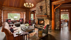 A log cabin interior with a stone fireplace, wooden furniture, and a large window overlooking a forest. - Fairmont Jasper Park Lodge (Jasper)
