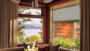 A cozy room with a large window offering a view of a lake and mountains. There's a round table with a book and a vase of flowers. - Fairmont Jasper Park Lodge (Jasper)