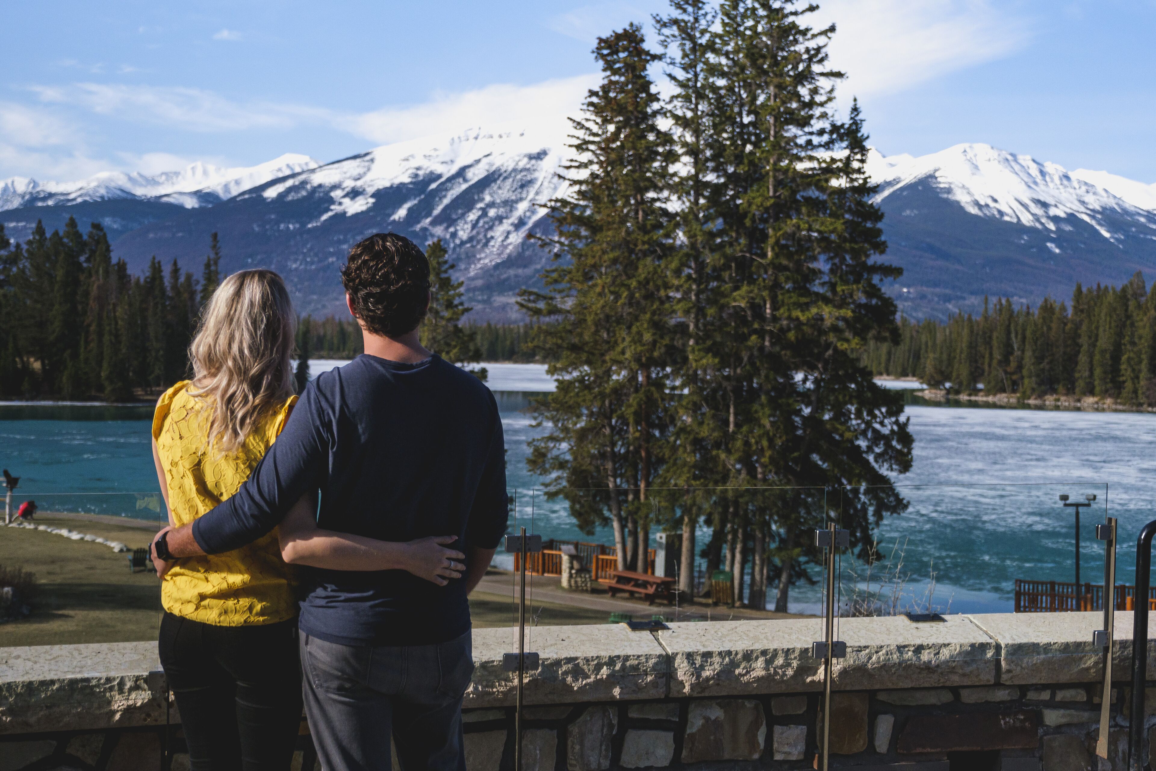 Un couple se tient au bord d’un lac, avec des montagnes en arrière-plan.