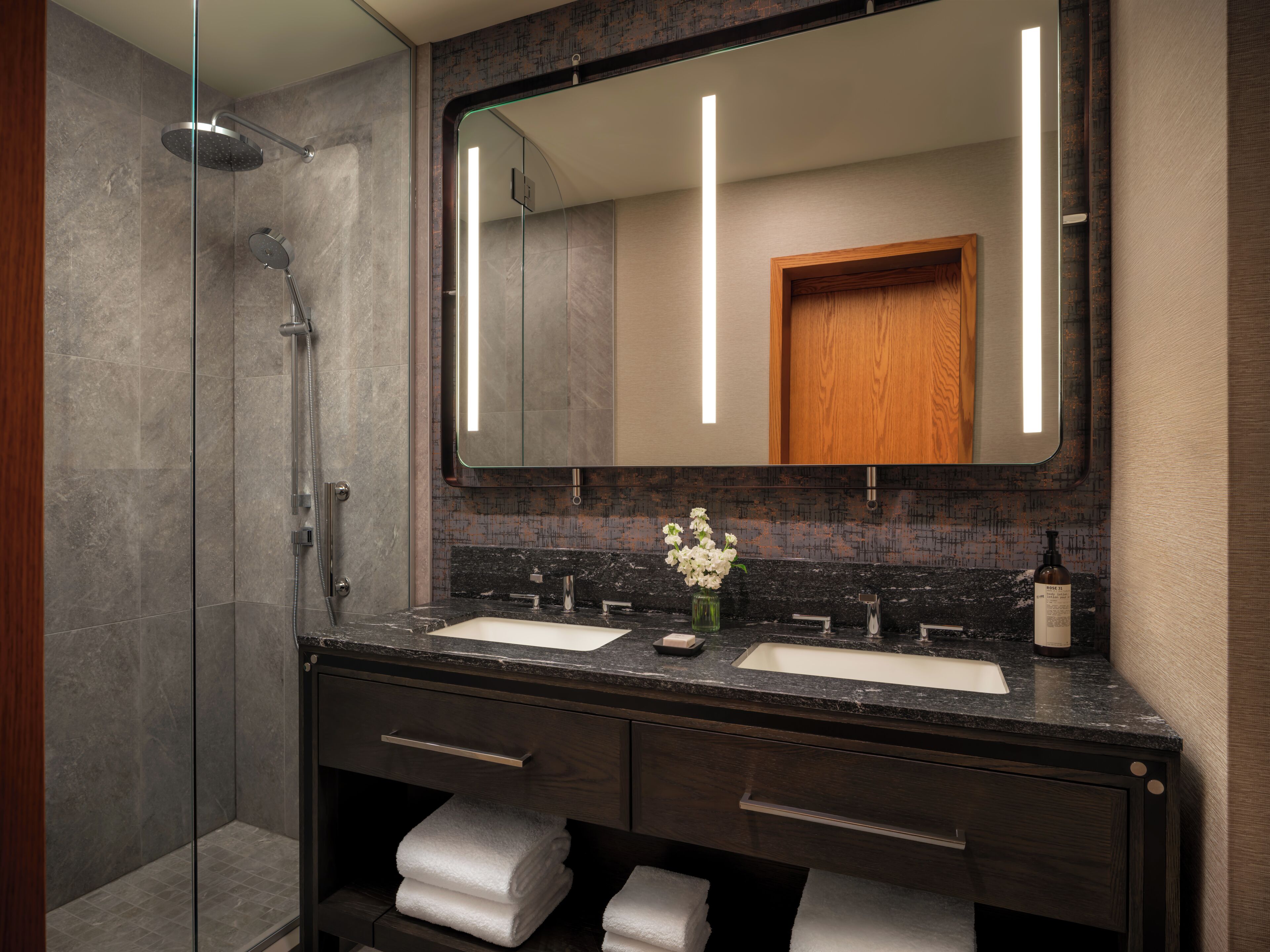 Un baño moderno con ducha de vidrio, dos lavamanos y un espejo grande.