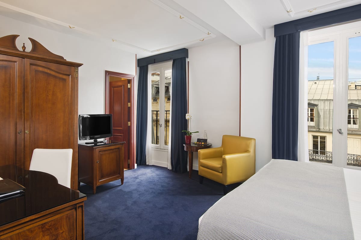 A hotel room with a bed, a desk with a television, a chair, a wardrobe, and a window with a view of buildings.