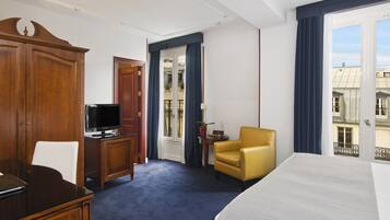 A hotel room with a bed, a desk with a television, a chair, a wardrobe, and a window with a view of buildings.