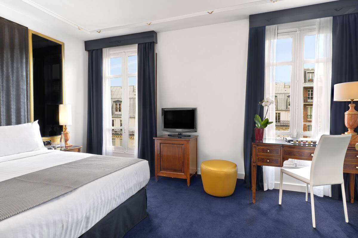 A hotel room with a large bed, a desk with a chair, a television, and a view of buildings through the windows.