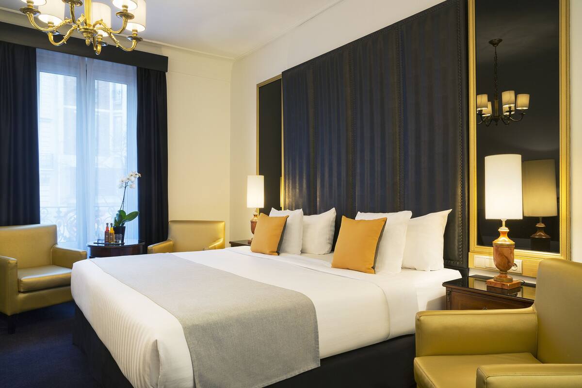 A hotel room with a large bed, two bedside tables, a chandelier, a potted plant, and a view of the city.