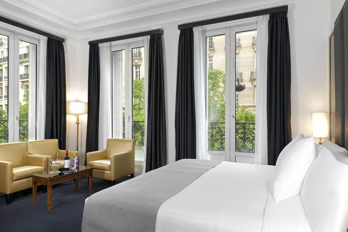 A hotel room with a large bed, two armchairs, a small table with a bottle and glasses, and a view of a building and greenery through the windows.