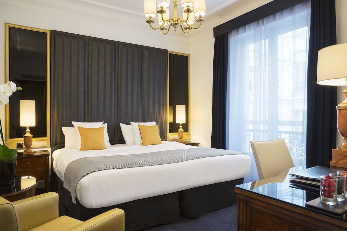 A hotel room with a large bed, bedside tables with lamps, a desk with a chair, and a large window with curtains.