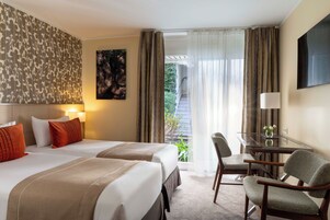 A hotel room with two beds, a desk with a chair, a TV, and a balcony with curtains.