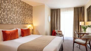 Quarto de hotel com uma cama grande, uma escrivaninha com cadeira, uma luminária e vista para o exterior através de cortinas.