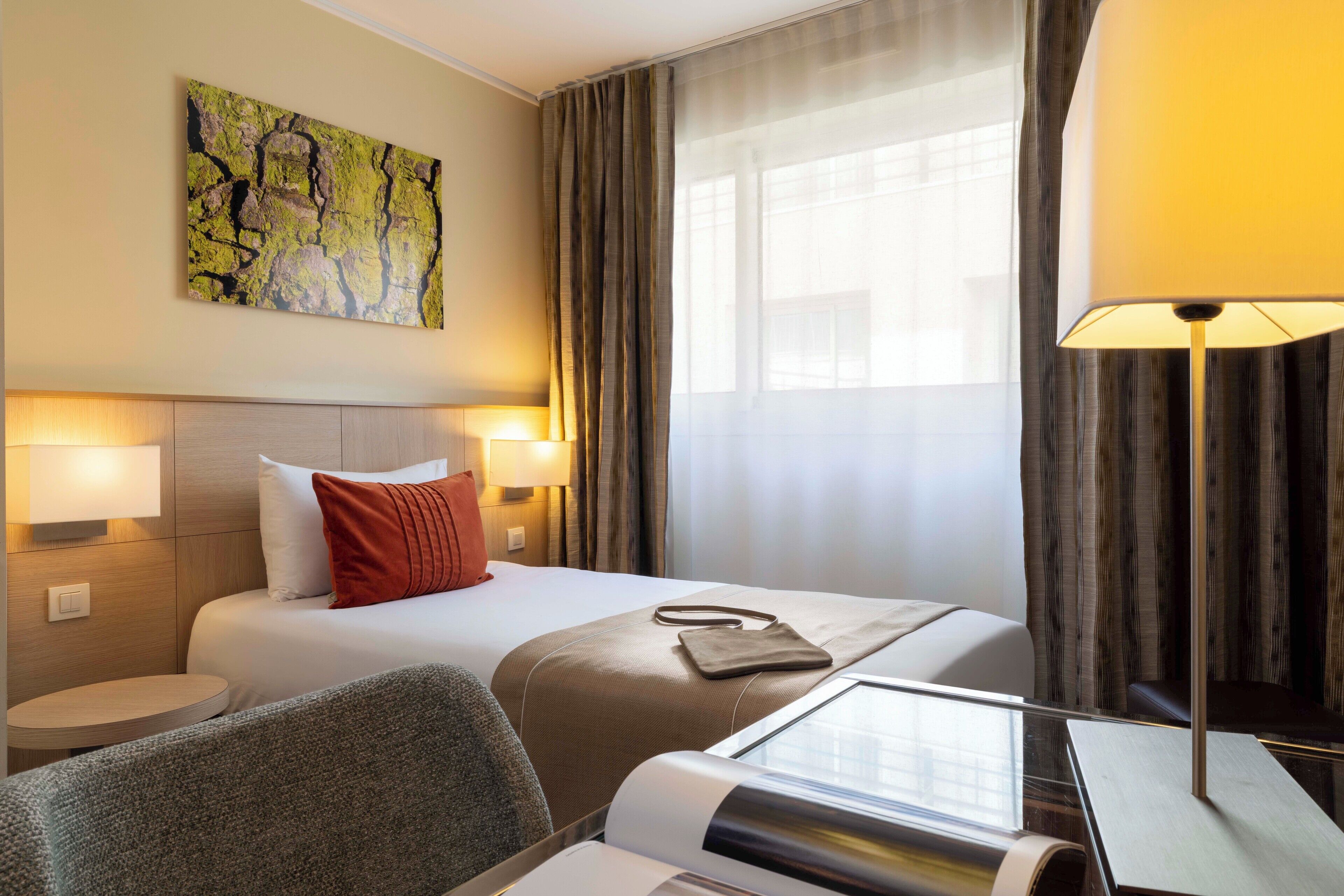 A hotel room with a bed, two bedside lamps, a painting on the wall, a window with curtains, and a desk with a lamp.