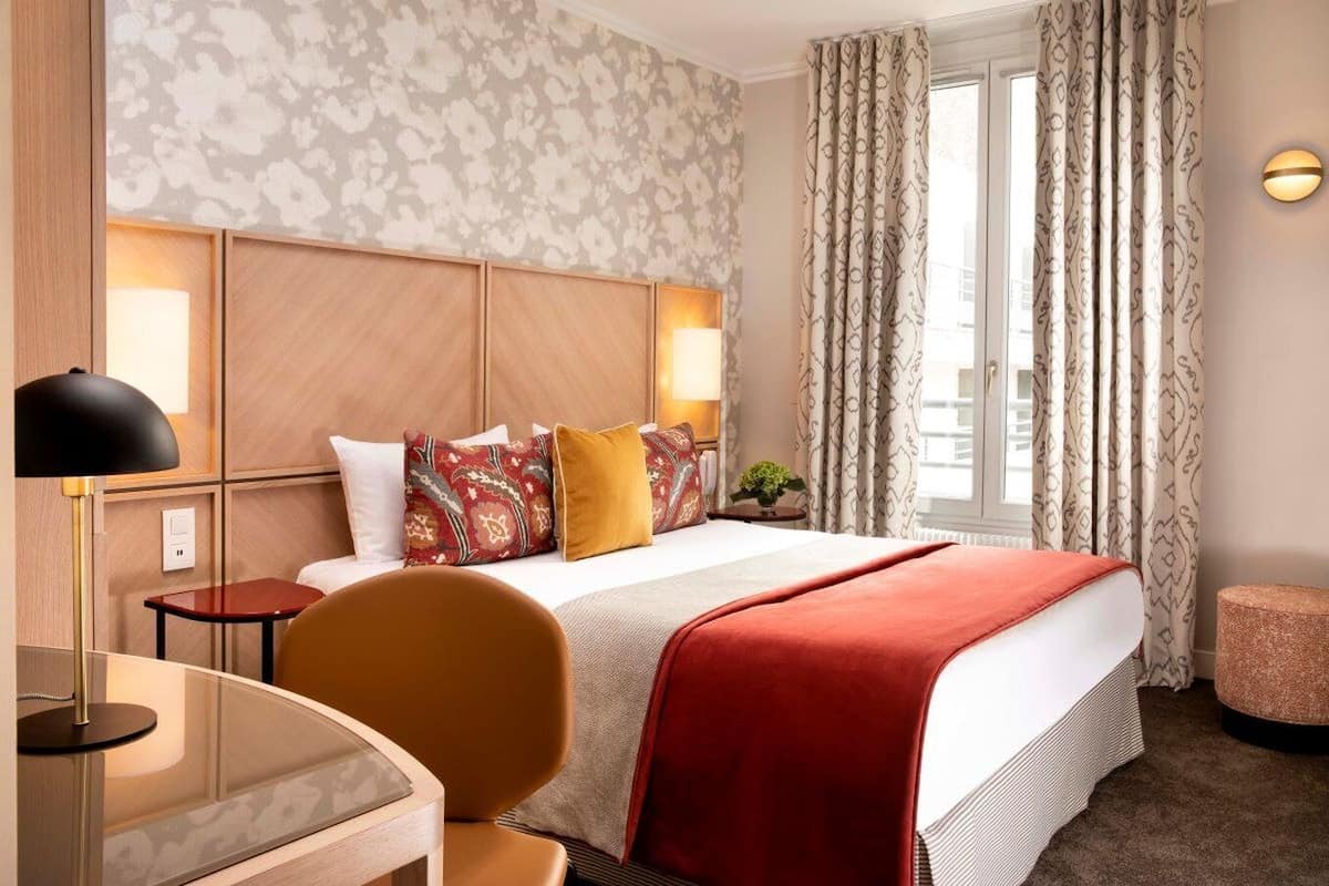 Ein Hotelzimmer mit Bett, Schreibtisch, Stuhl, Lampe und einem Vorhangfenster.