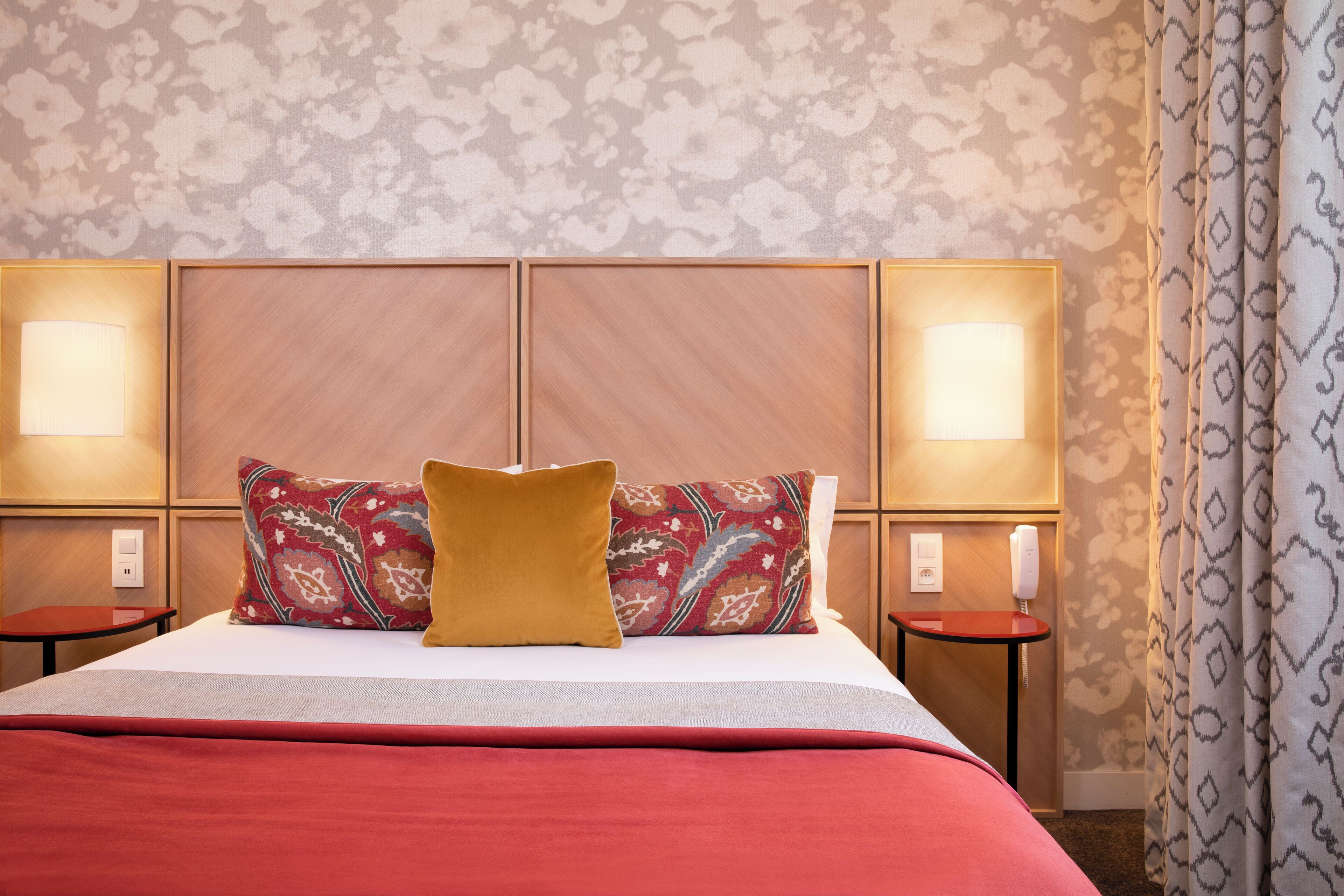 A hotel room with a large bed, two bedside tables, a patterned wall, and a patterned curtain.