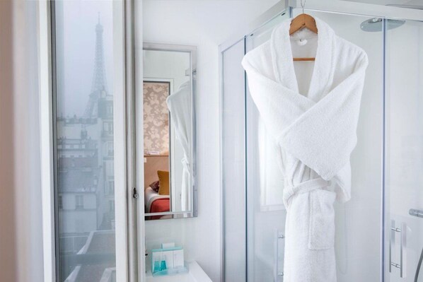 A hotel bathroom with a glass shower enclosure, a white robe hanging on a rack, and a view of the Eiffel Tower through a window. - Les Jardins d'Eiffel (Paris)