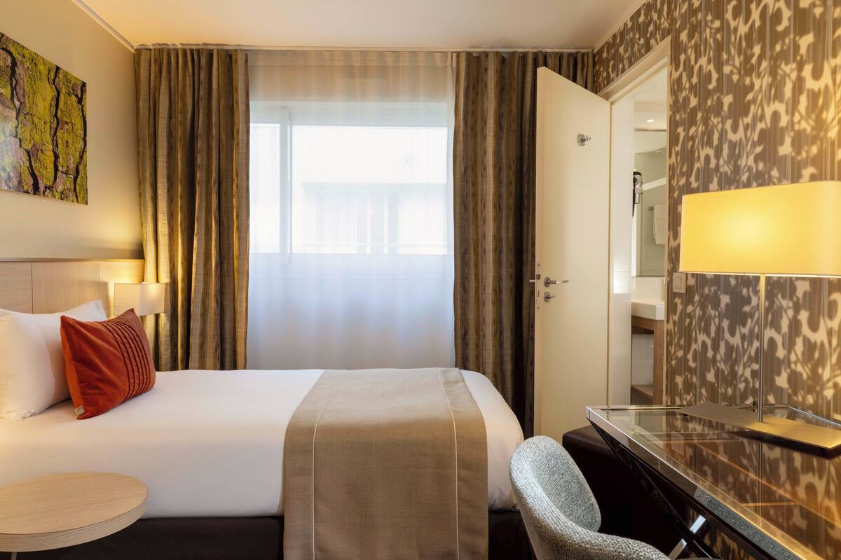 Ein Hotelzimmer mit einem Bett, einem Schreibtisch, einem Sessel, einem runden Tisch, einem großen Fenster mit Vorhängen, einem Bild an der Wand und einem Spiegel.