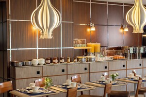 A restaurant with multiple tables, each set with white cups, saucers, and plates. There are decorative light fixtures above the tables, and a counter with various food items and condiments.