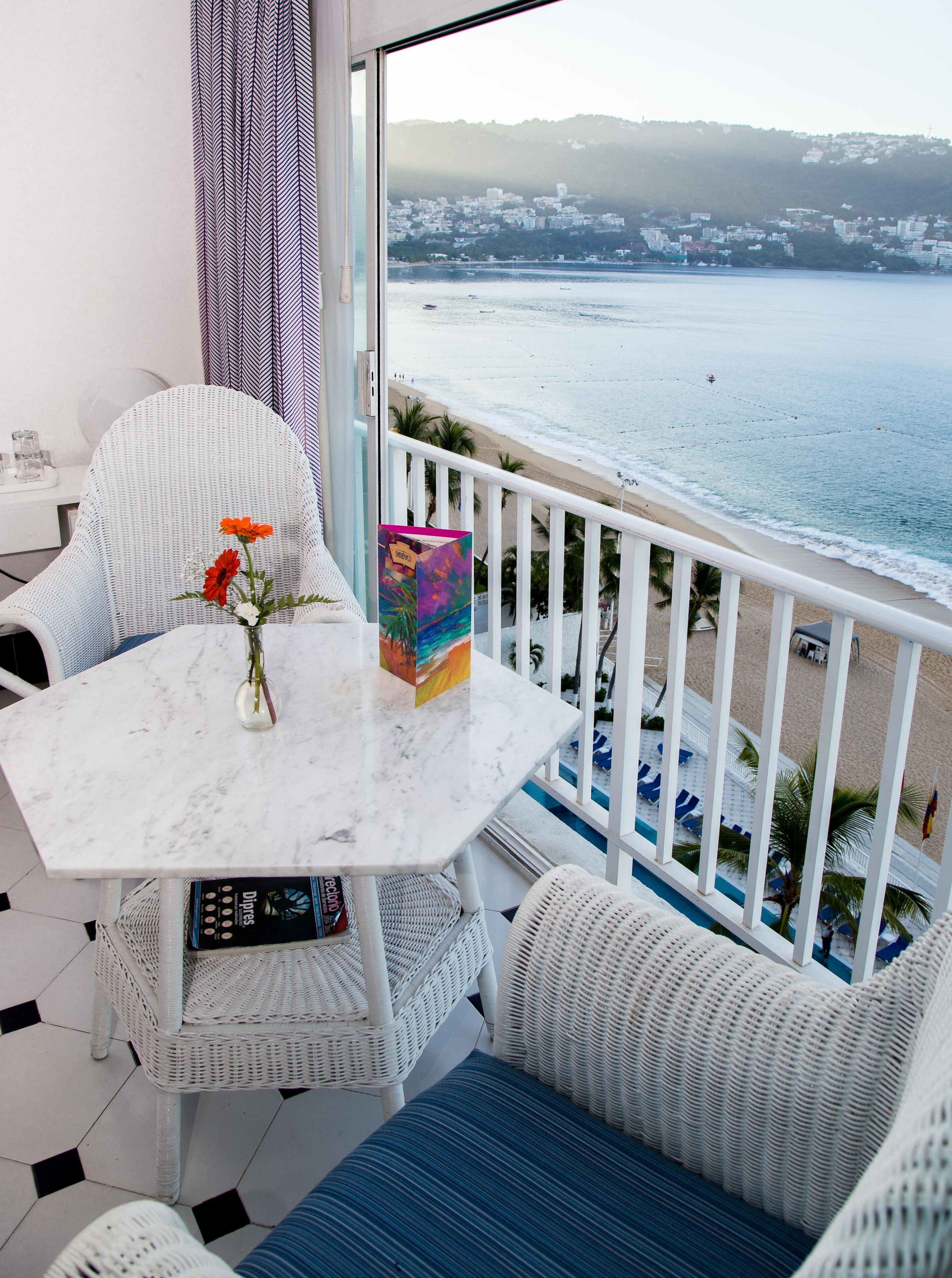 Un balcón con una silla de mimbre blanca, una mesita con un florero y un libro colorido. El balcón da a una playa con vista al mar y edificios distantes.