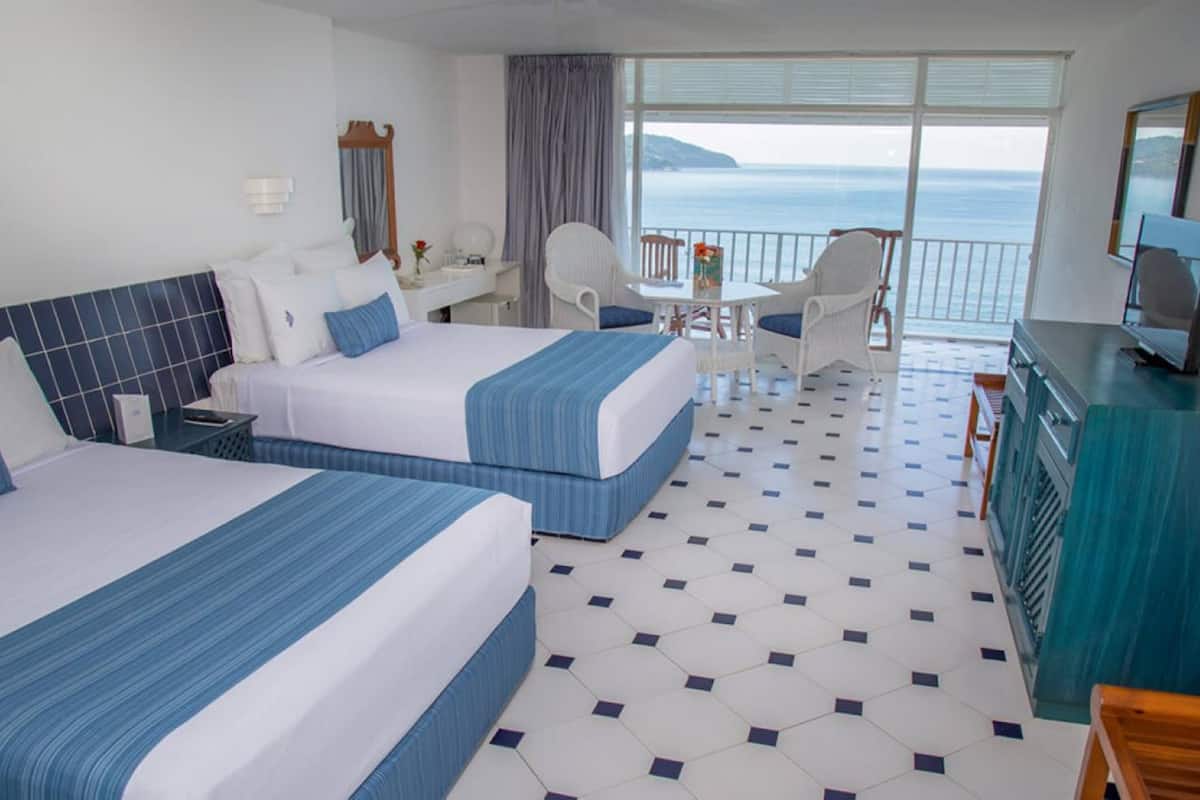 A hotel room with two beds, a balcony with a view of the sea, a desk, and a television.