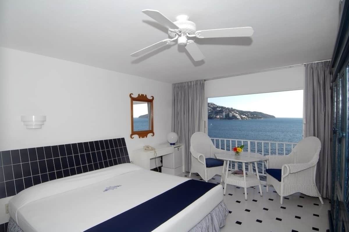 A hotel room with a bed, a desk, a chair, a table with flowers, a ceiling fan, a view of the sea, and a balcony with curtains.