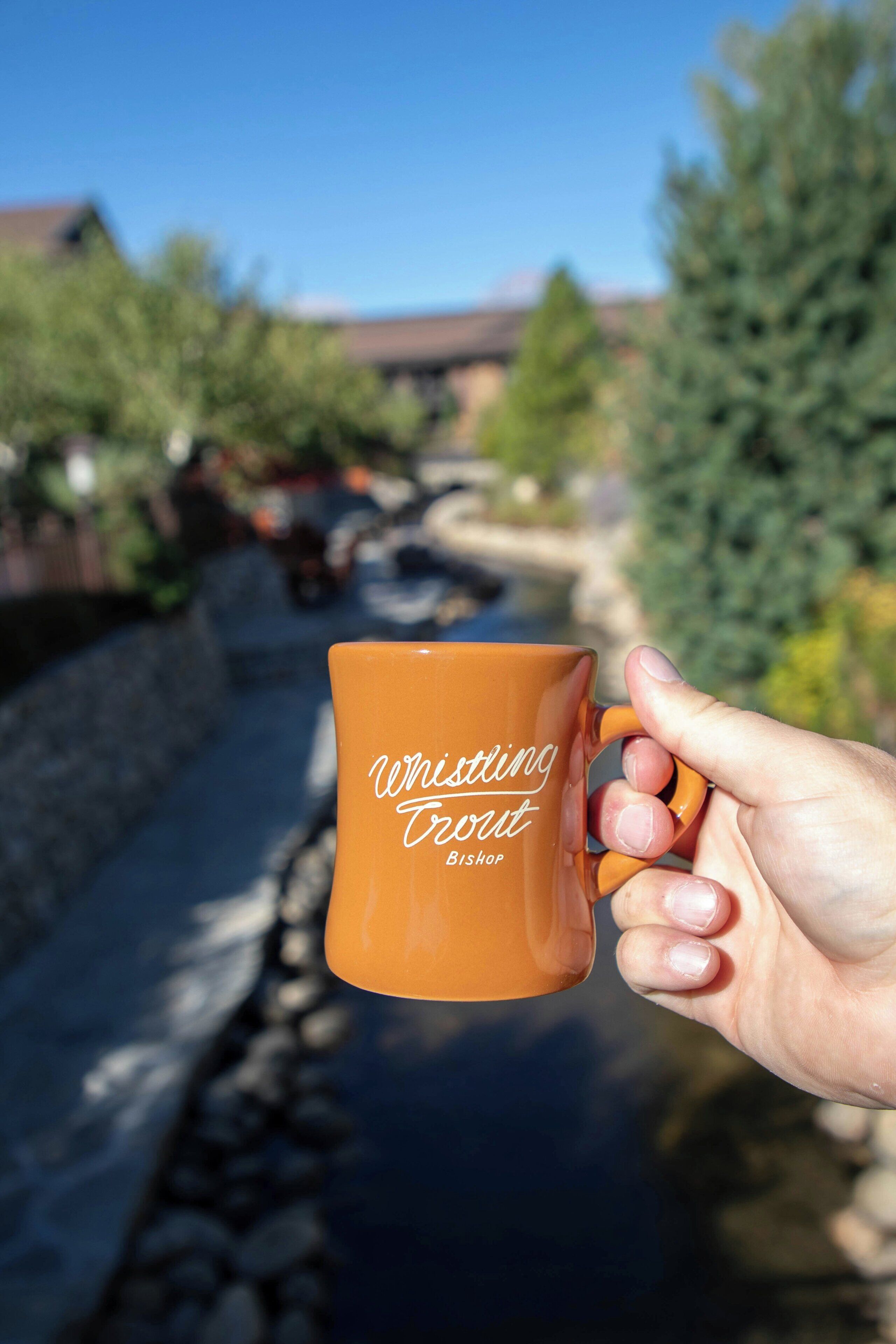 A hand holding a mug with the inscription 'Whistling Trout Bishop.'
