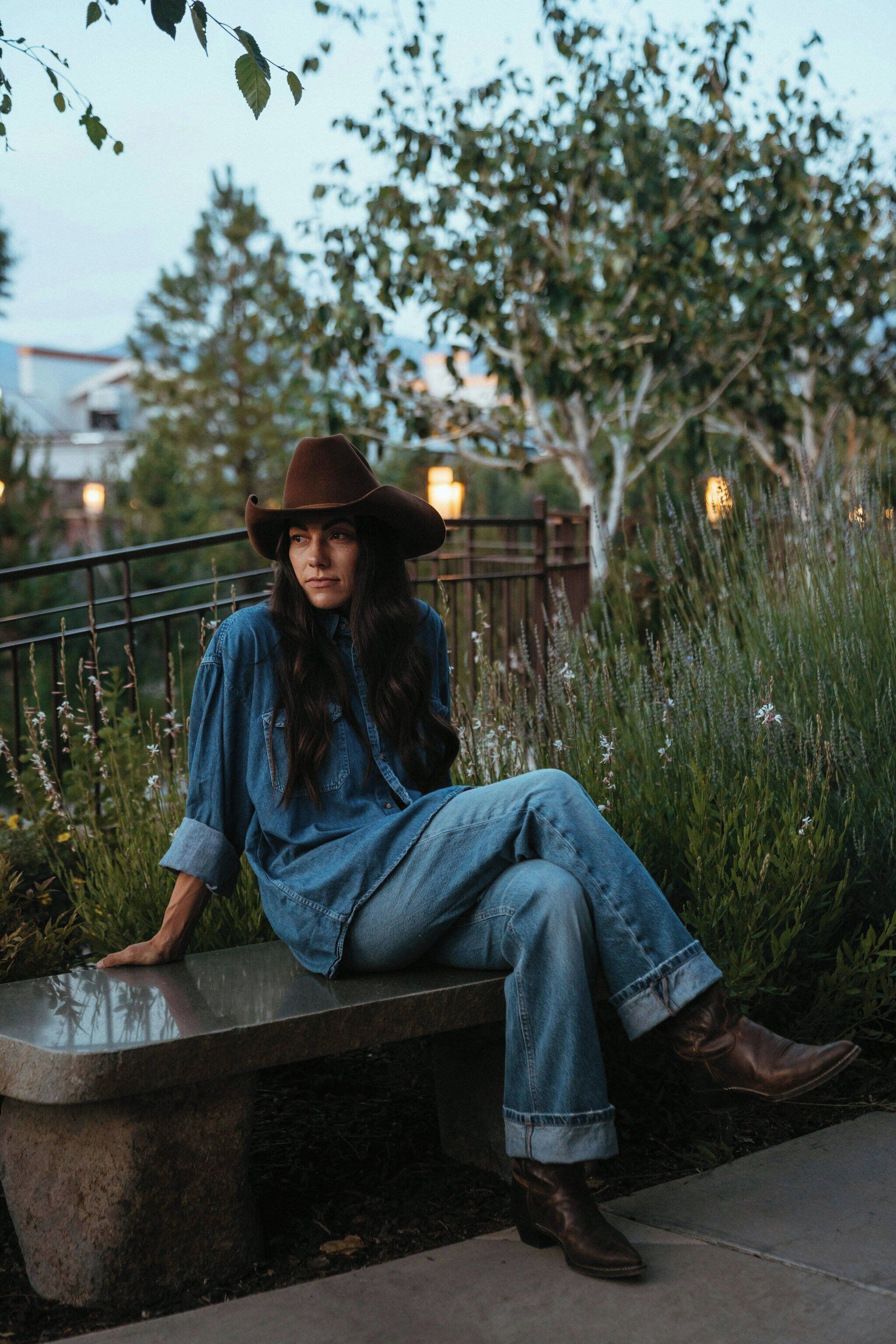 A person sitting on a bench outdoors with greenery and a fence in the background.