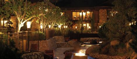 A stone pathway leading to a building with a fire feature, surrounded by trees and a small stream.