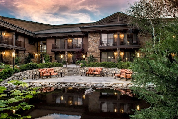 A two-story building with a stone facade, outdoor seating area with chairs, and a small pond. - Wayfinder Bishop (Bishop)