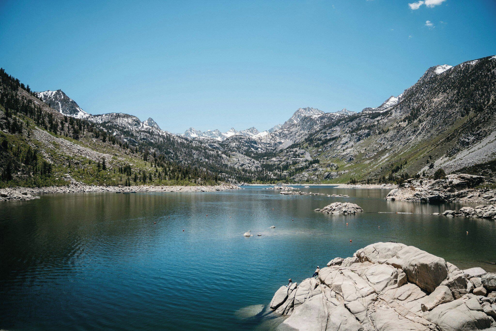 A mountain lake with clear water, surrounded by rocky terrain and snow-capped peaks.