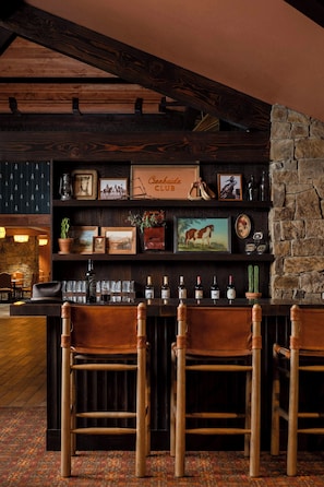 Una zona de bar con sillas de madera, una pared de piedra y varias fotografías enmarcadas y botellas en estanterías.