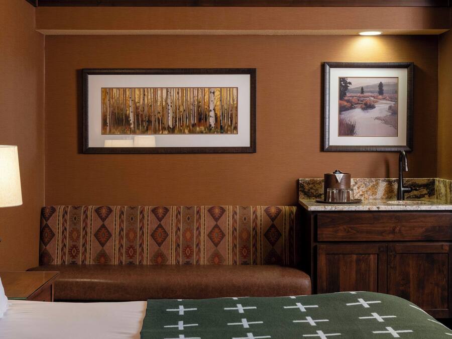 A hotel room with a bed, a bench, a small table with a lamp, and two framed pictures on the wall.