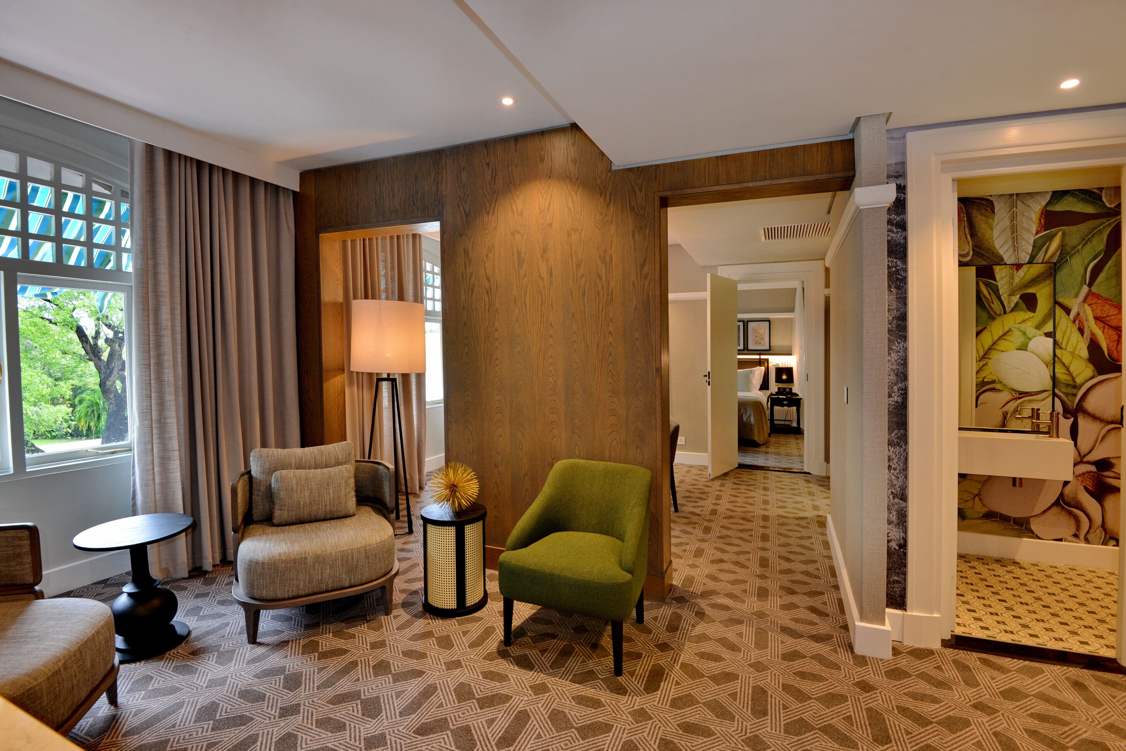 A hotel room with a patterned carpet, a green armchair, a beige armchair, a small round table, and a lamp.