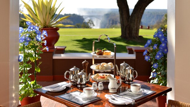 A table set for a tea service with a scenic view of a bridge and greenery. - The Victoria Falls Hotel (Victoria Falls)
