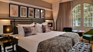 A neatly arranged hotel room with a large bed, a side table, a green armchair, and a window with curtains. - The Victoria Falls Hotel (Victoria Falls)