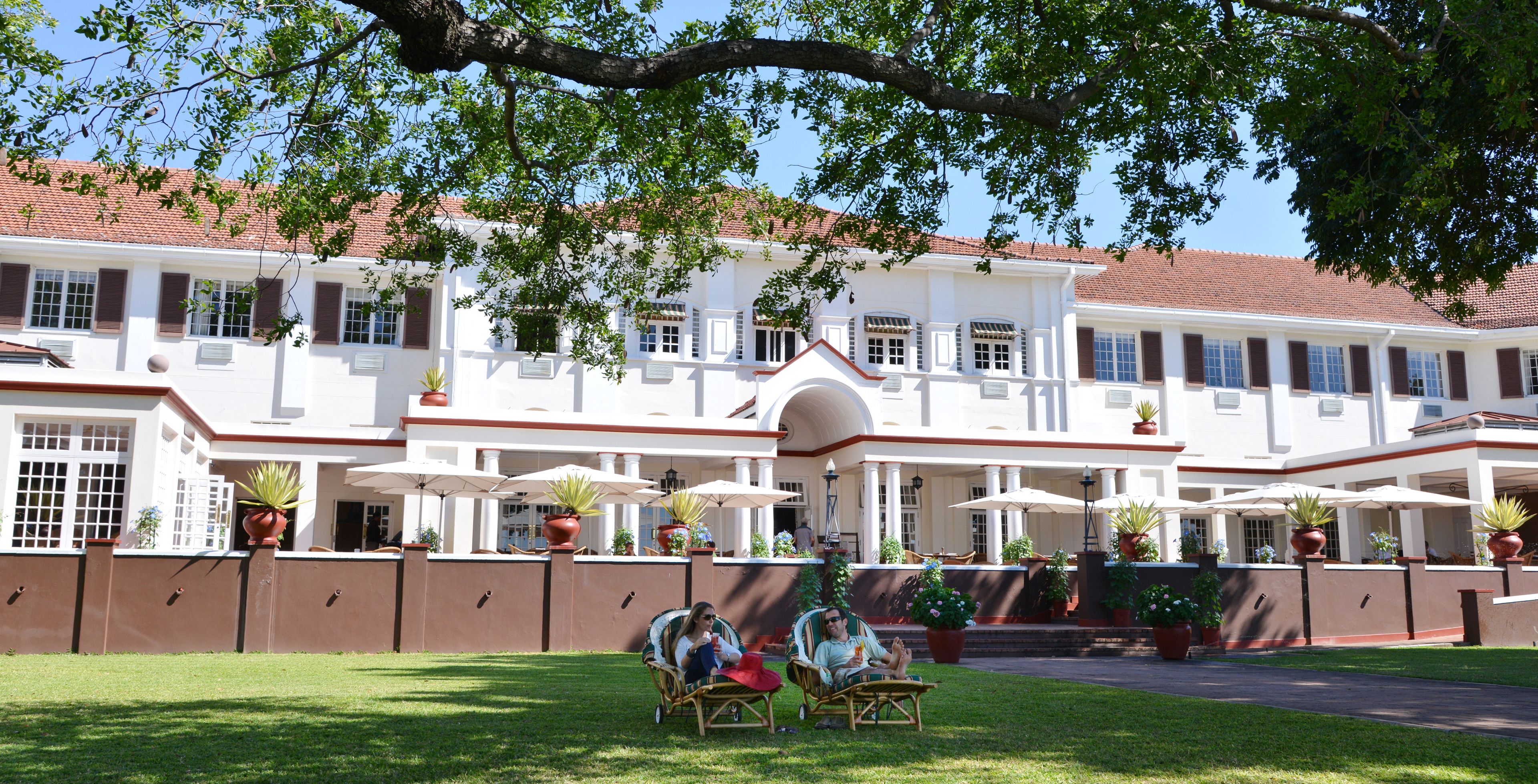 A large white building with a red-tiled roof, multiple windows, and outdoor seating areas with umbrellas.