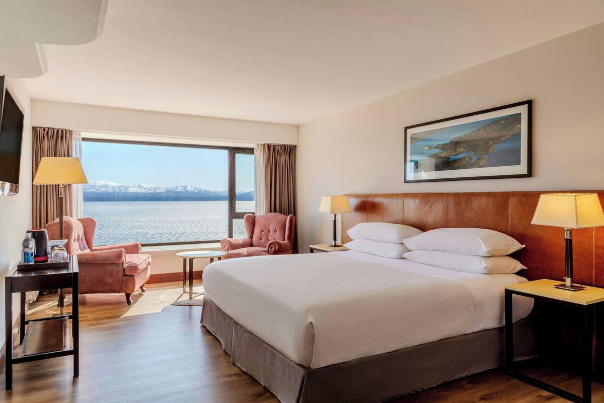 Une chambre d’hôtel avec un grand lit, deux tables de chevet, une télévision, un bureau avec une lampe, une chaise et une vue sur la mer.