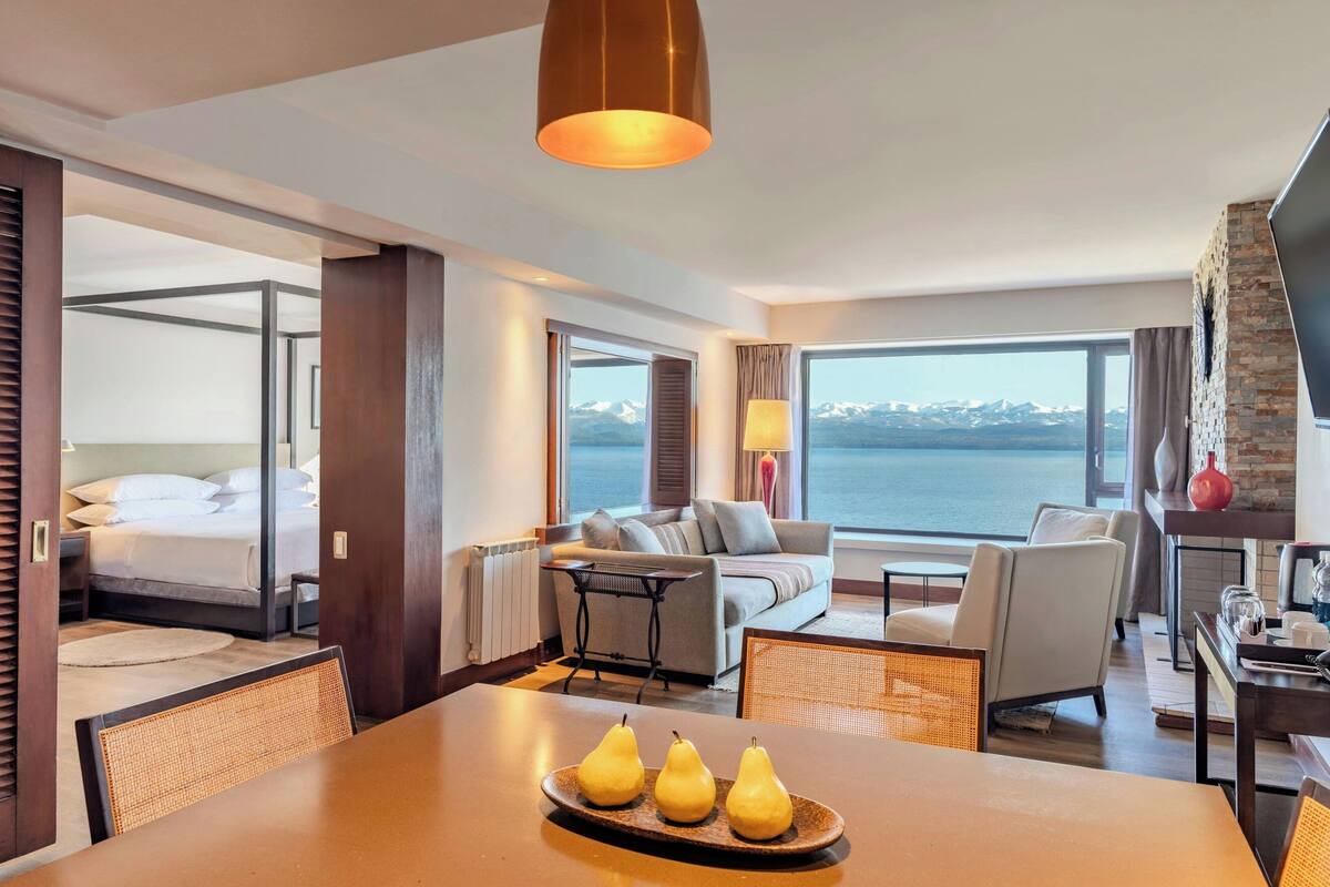 Une chambre d’hôtel avec un lit, un canapé, une table à manger avec des poires, et une vue sur la mer.