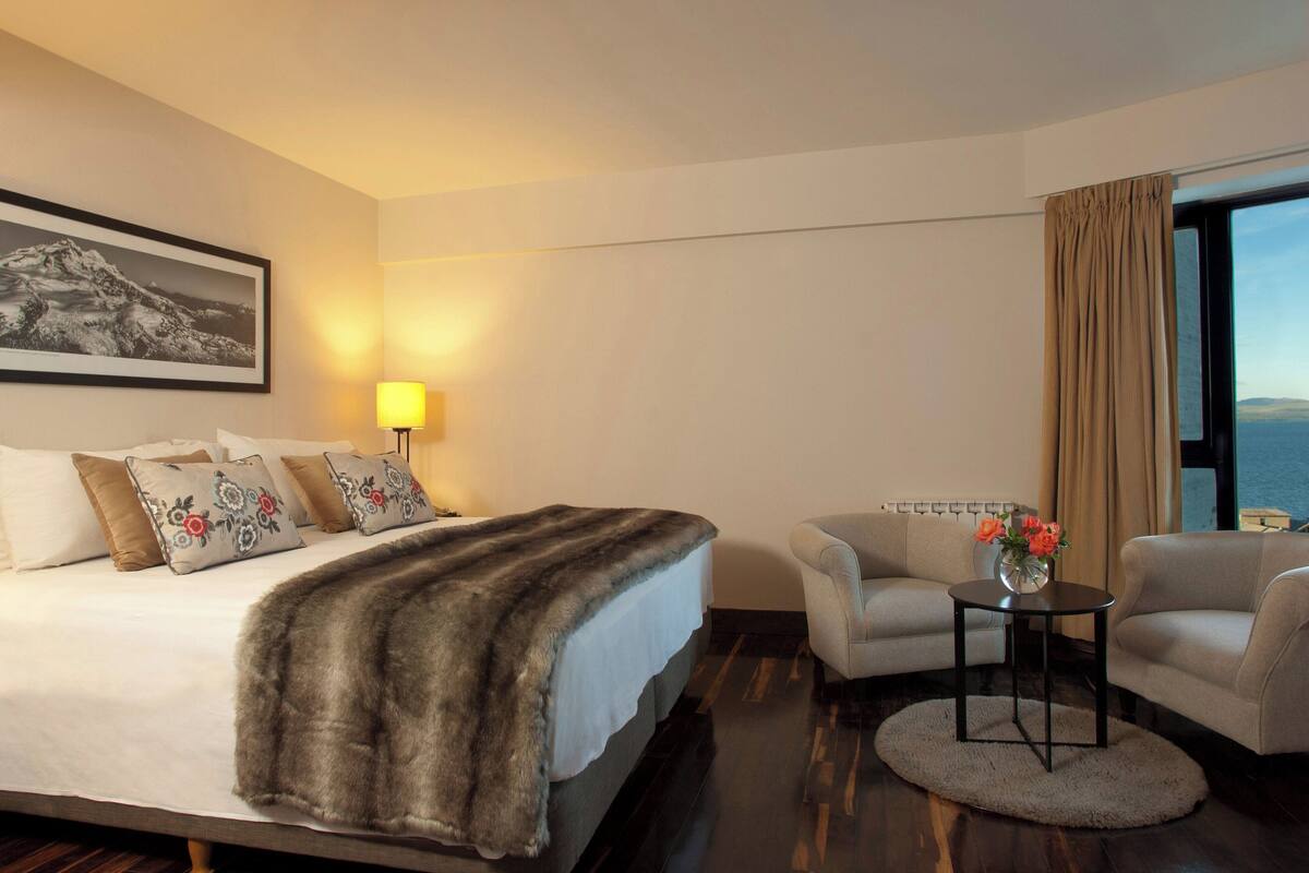 Une chambre d’hôtel avec un lit, deux fauteuils, une petite table sur laquelle est posé un vase de fleurs, une lampe et une grande fenêtre avec des rideaux.