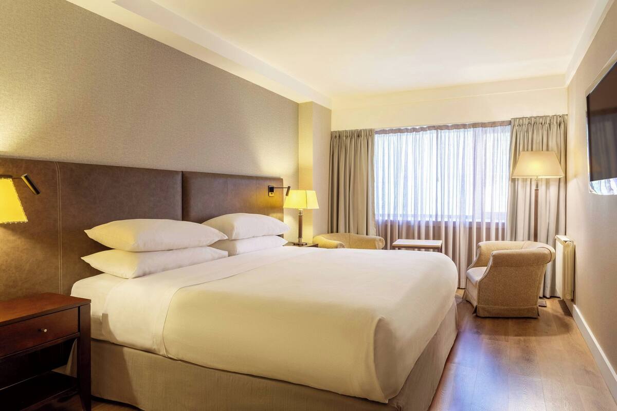 Une chambre d’hôtel avec un grand lit, deux tables de chevet, une chaise et une fenêtre avec des rideaux.
