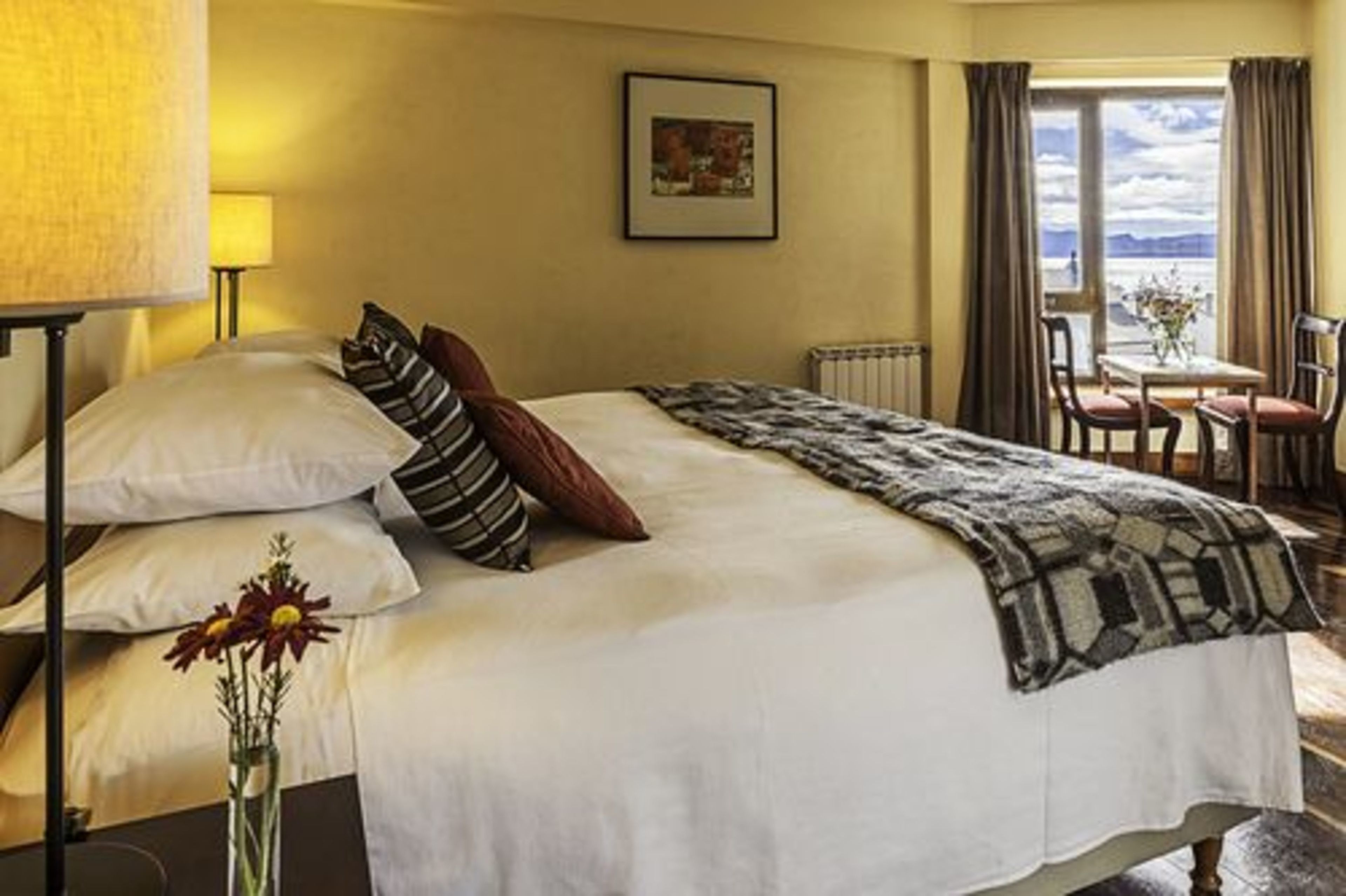 Habitación de hotel con cama, una mesa con sillas, una ventana con cortinas y un cuadro en la pared.