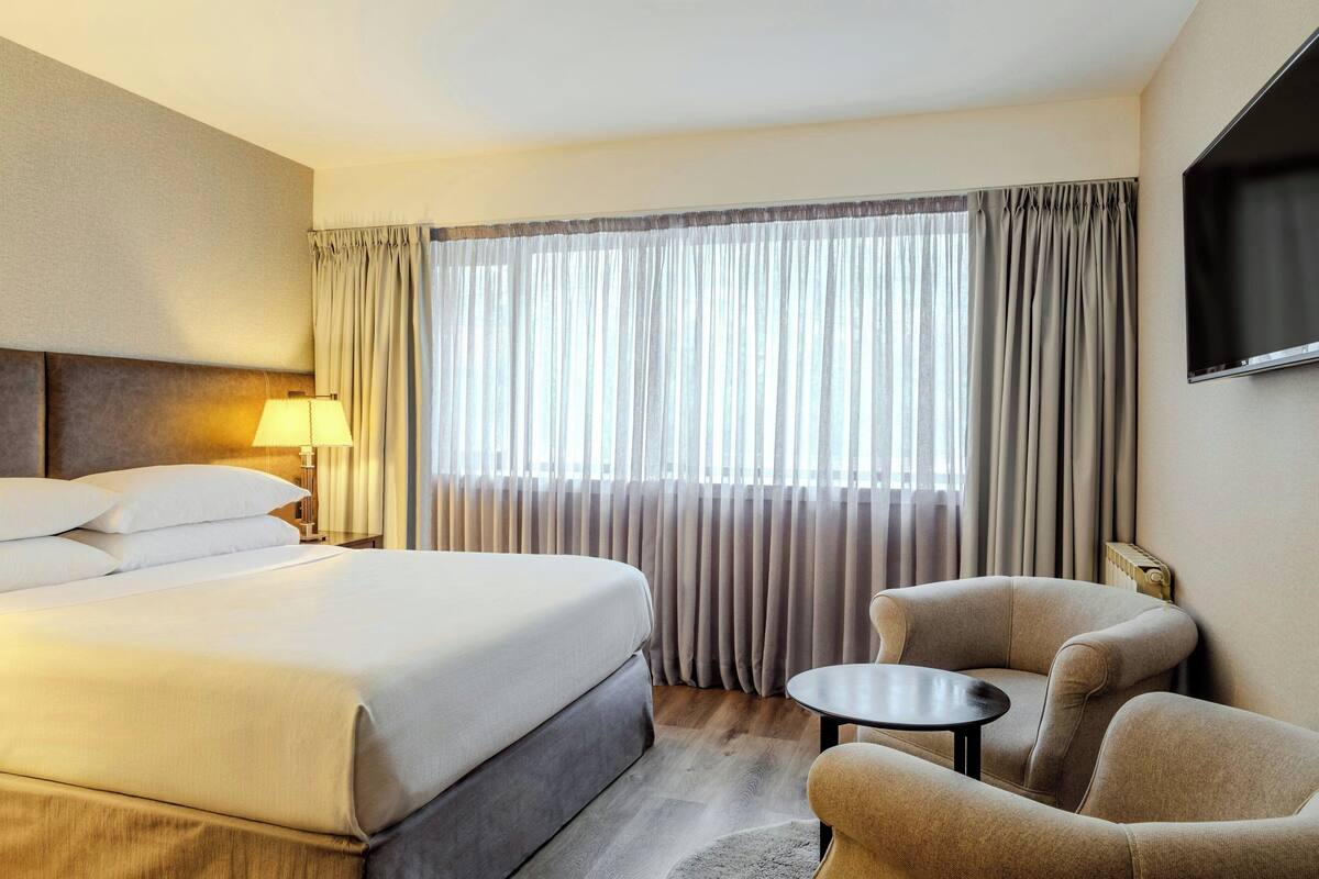 Une chambre d’hôtel avec un lit, deux fauteuils, une petite table, un téléviseur et une fenêtre avec des rideaux.