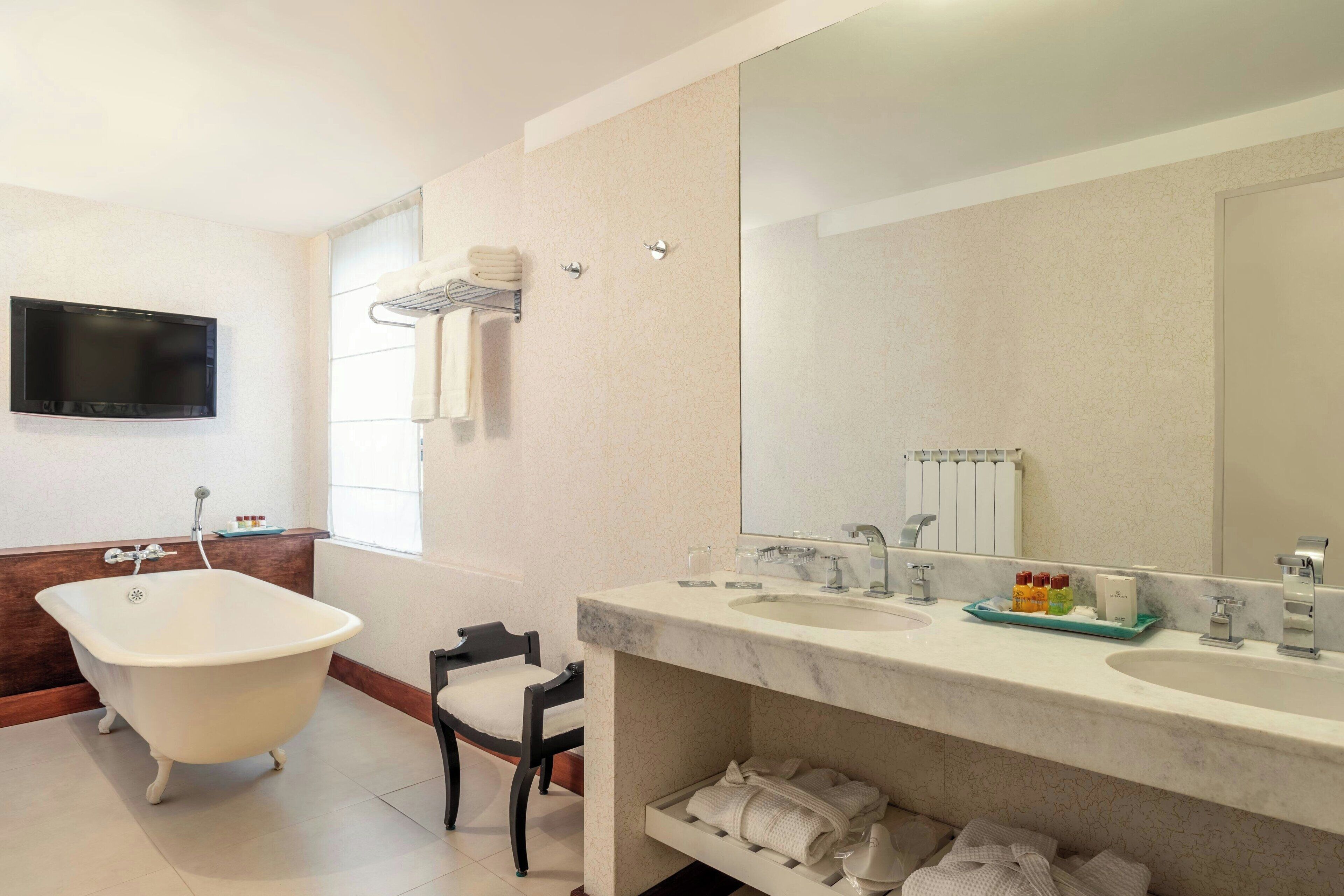 A bathroom with a freestanding bathtub, two sinks, a large mirror, and a television mounted on the wall.