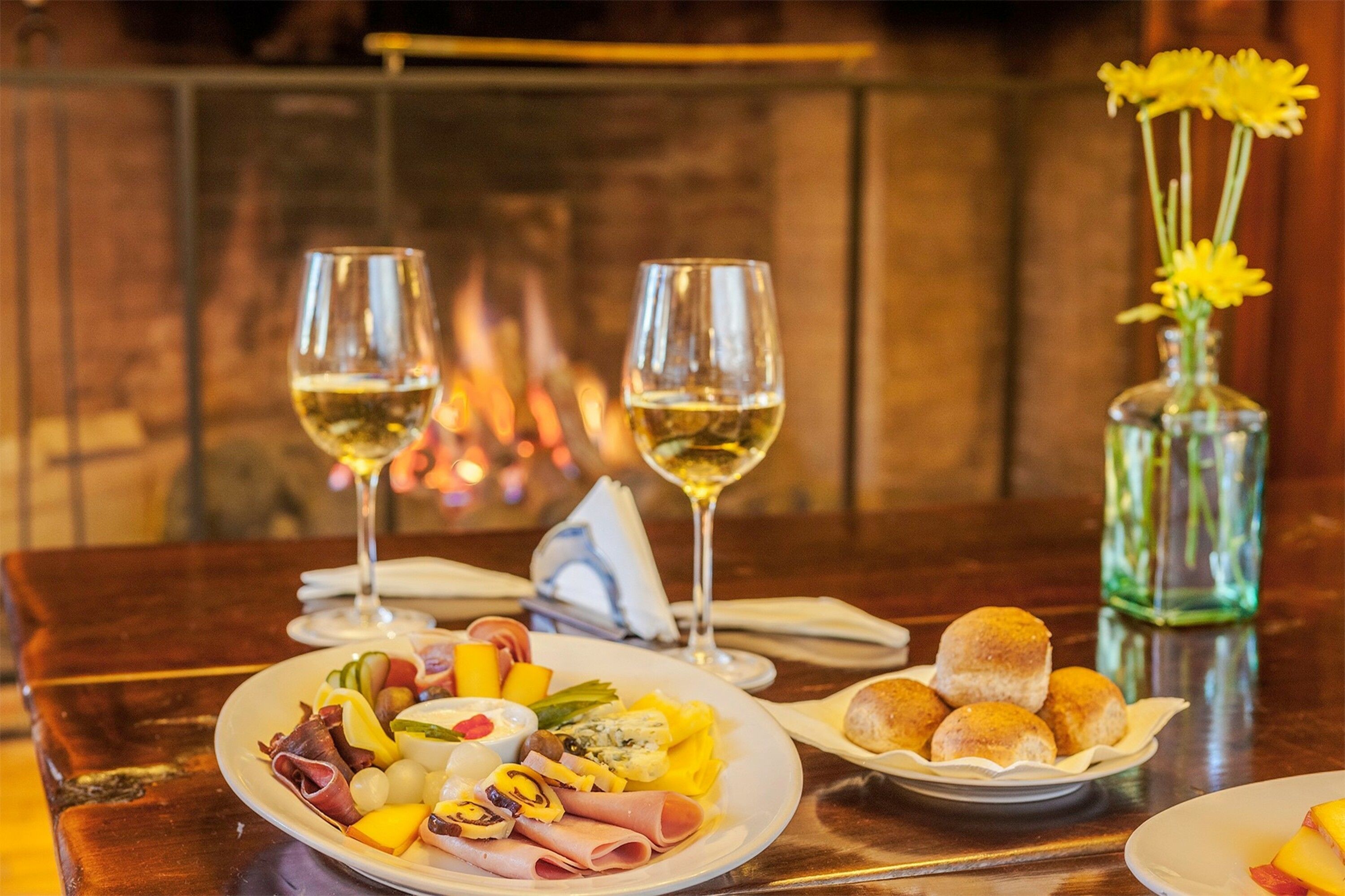 A dining table with a charcuterie board, two glasses of white wine, a vase with yellow flowers, and a plate of bread rolls.