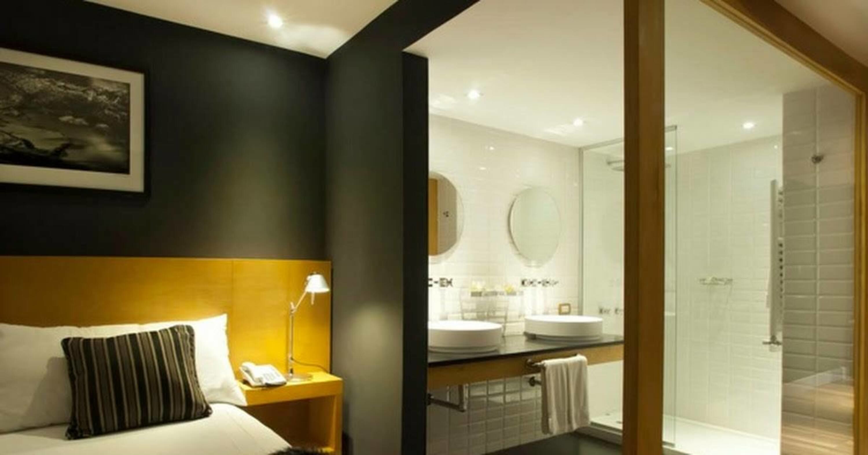 Une chambre d’hôtel avec un lit, une table de chevet avec une lampe, un téléphone, un tableau encadré au mur, une salle de bain avec deux lavabos, un miroir et une douche en verre.