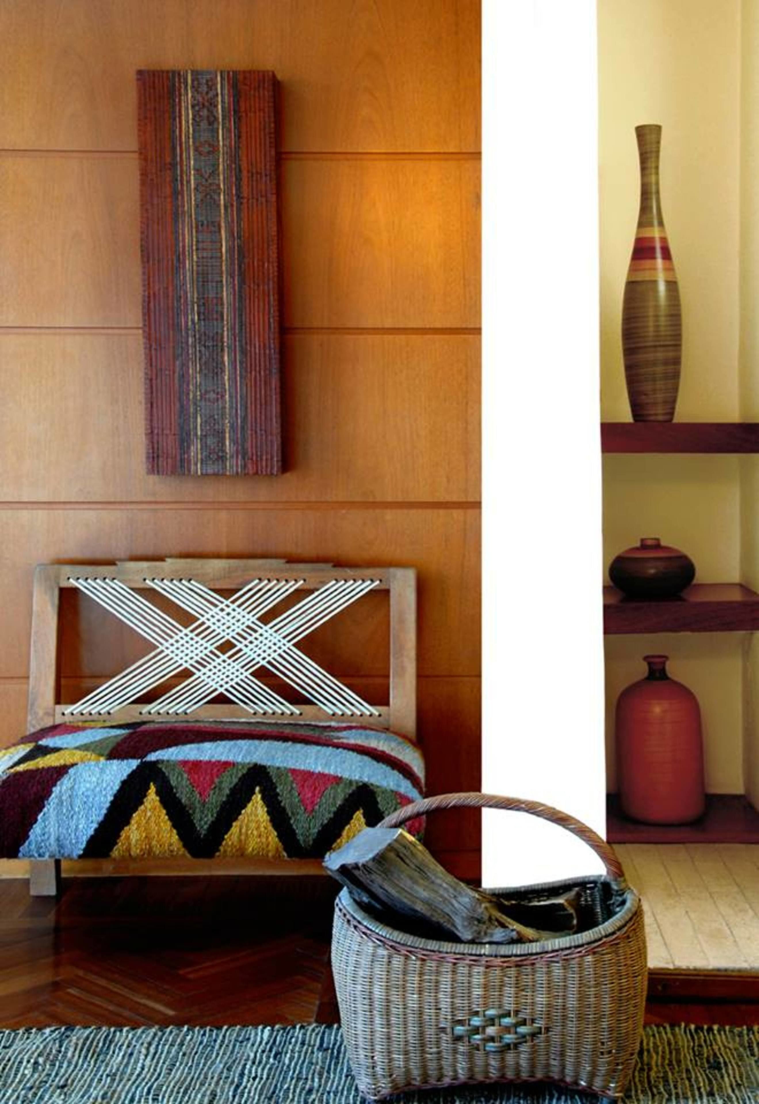 A room with a wooden headboard, a colorful woven bedspread, a woven basket, and a shelf with decorative items.