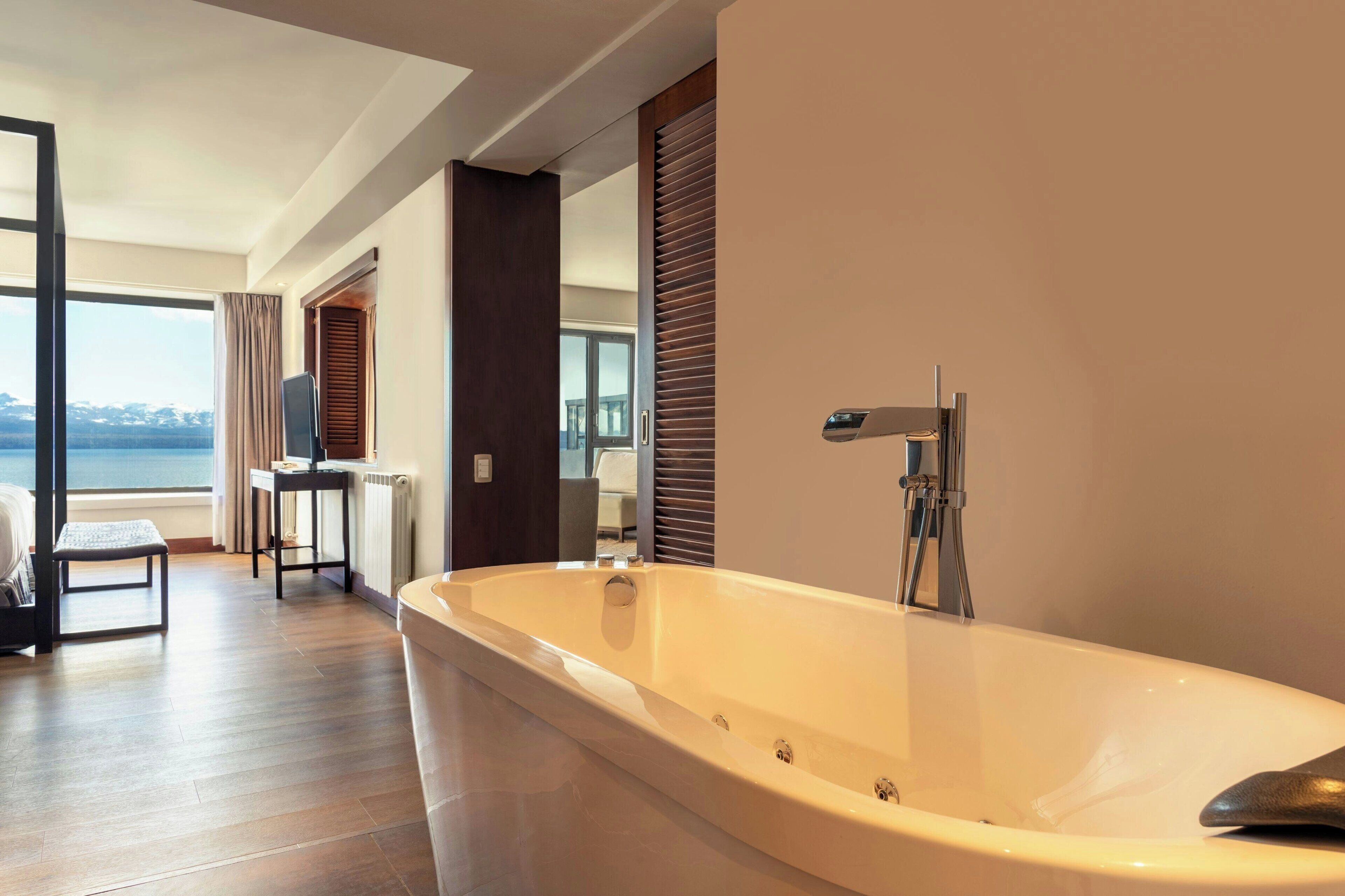 A modern bathroom with a large bathtub, wooden flooring, and a view of the sea through a large window.