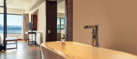 A modern bathroom with a large bathtub, wooden flooring, and a view of the sea through a large window.