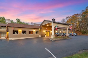 A hotel with a parking lot, multiple flags, and a sunset sky. - Skyline Hotel Lake George (Lake George)