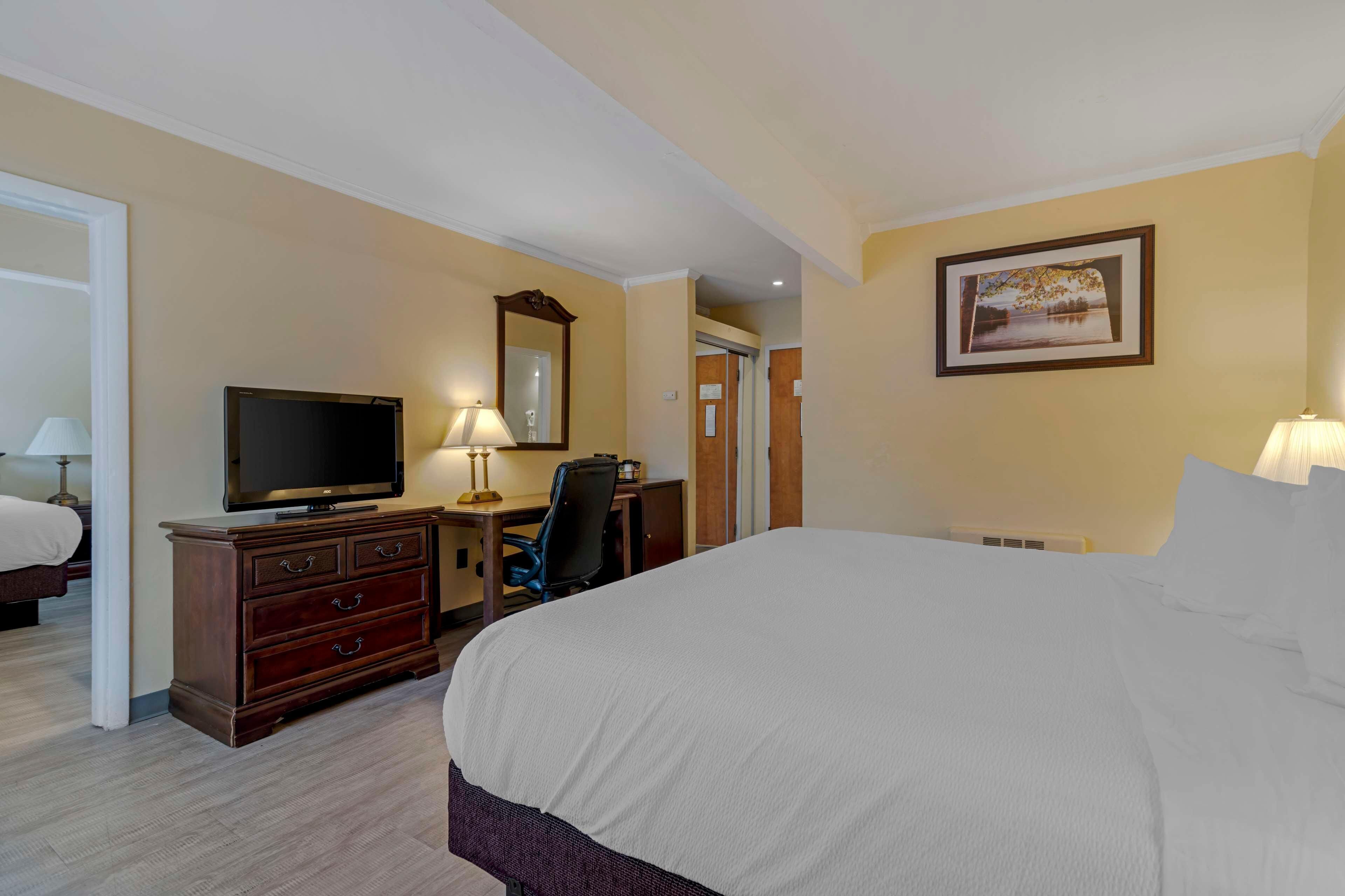 Habitación de hotel con una cama, un escritorio con televisión, una silla, una lámpara de escritorio, un espejo y un cuadro en la pared.