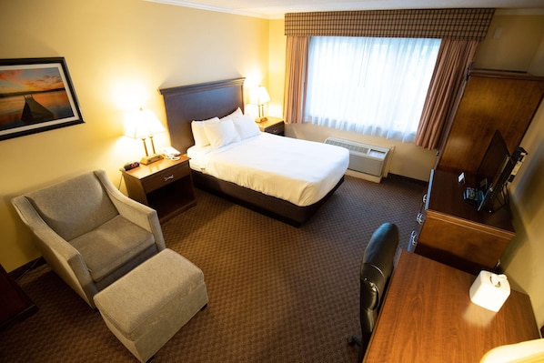 A hotel room with a bed, a chair, a desk, a lamp, a clock, and a window with curtains.